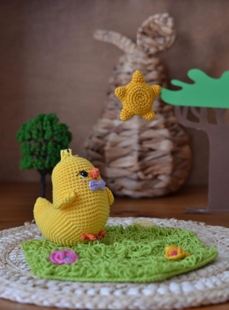 Puede incluir: Un pollito amarillo de ganchillo hecho a mano con una pajarita morada se sienta sobre una base verde de ganchillo con una flor y una estrella amarilla. Un conejo tejido y un peque&ntilde;o &aacute;rbol est&aacute;n en el fondo. Decoraci&oacute;n caprichosa.