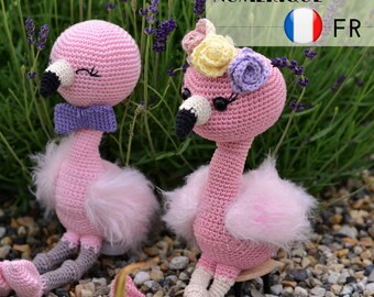 May include: Two pink crocheted flamingo toys with fluffy pink wings. One flamingo has a purple bow and the other has a flower crown with pink, yellow, and purple flowers.
