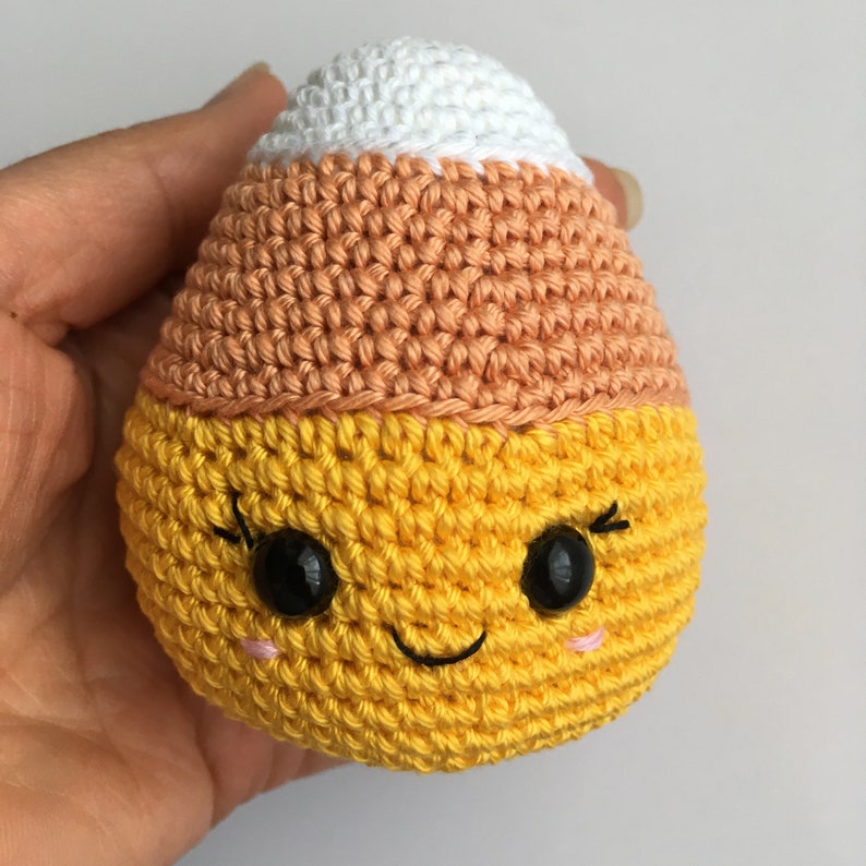 May include: A crocheted candy corn toy with a yellow body, orange middle section, and white top. The toy has a smiling face with black eyes and a black mouth.
