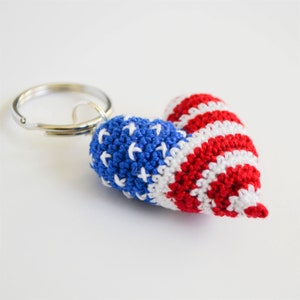 May include: A crocheted heart-shaped keychain in the colors of the American flag. The keychain is red, white, and blue and features a star pattern on one side and stripes on the other.