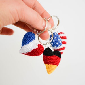 May include: Three crocheted heart-shaped keychains with the flags of the United States, France, and Germany. The keychains are attached to silver key rings.