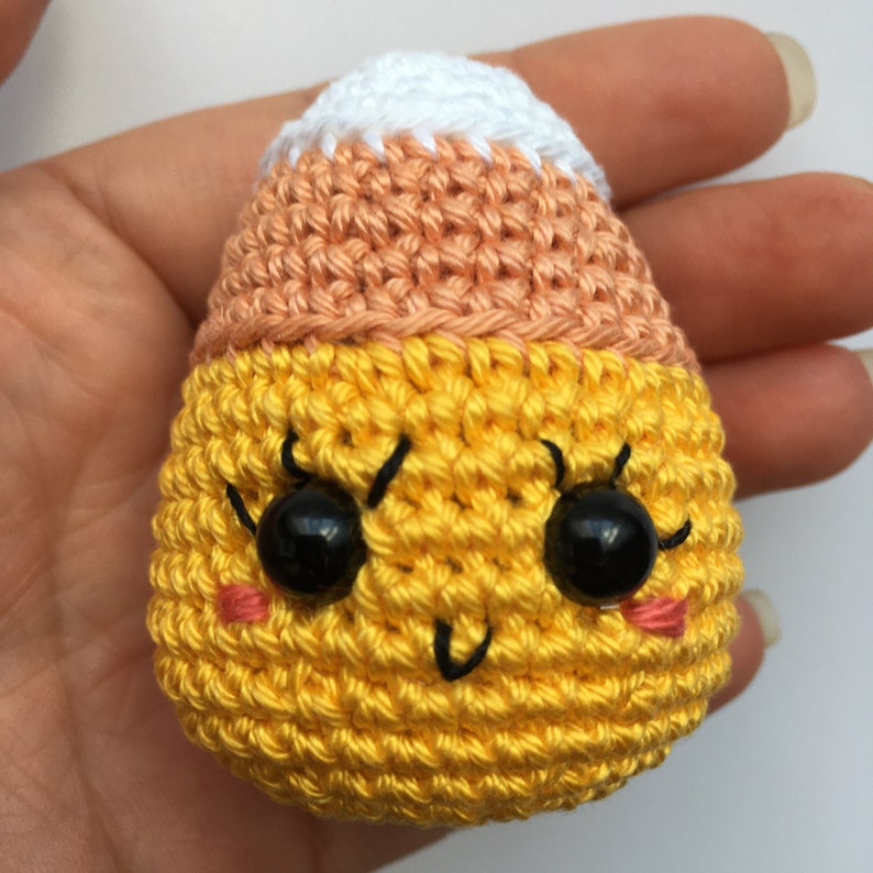 May include: A crocheted candy corn with a yellow body, orange top, and white tip. The candy corn has a cute face with black eyes, a black smile, and pink cheeks.