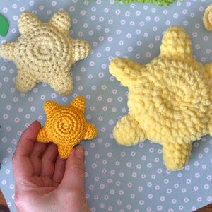 Puede incluir: Estrellas de mar de ganchillo hechas a mano en tonos amarillos. Una peque&ntilde;a estrella de mar se sostiene en la mano, mientras que dos estrellas de mar m&aacute;s grandes se exhiben cerca. Fondo azul claro con lunares blancos.