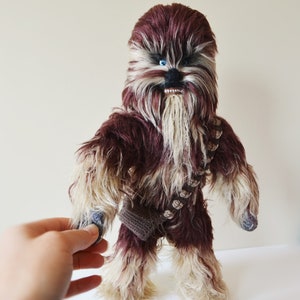 May include: A brown and white furry Chewbacca plush toy with blue eyes. The toy is wearing a brown and white bandolier.