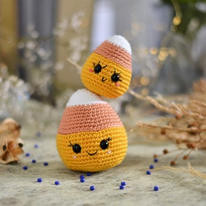 May include: Two crocheted candy corn plushies with smiling faces. The plushies are stacked on top of each other. The top plush is white and orange, and the bottom plush is white, orange, and yellow.