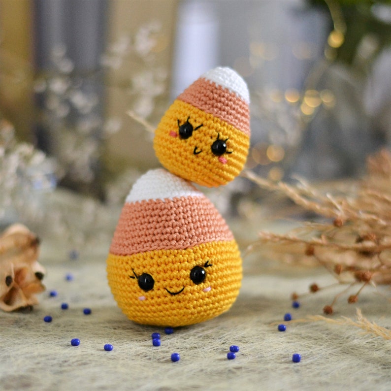 May include: Two crocheted candy corn plushies with smiling faces. The plushies are stacked on top of each other. The top plush is white and orange, and the bottom plush is white, orange, and yellow.