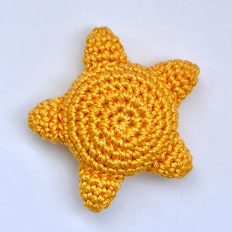 Puede incluir: Estrella de ganchillo dorada hecha a mano. La estrella tiene cinco puntas y un centro en espiral. El hilo tiene un ligero brillo, dando a la estrella una apariencia brillante. El art&iacute;culo artesanal es probablemente un elemento decorativo o un juguete.