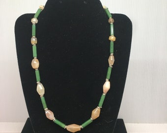 May include: A beaded necklace with a mix of green, tan, and silver-colored beads. The necklace is displayed on a black jewelry stand against a white background. The beads are various shapes, including cylindrical and oval.