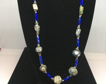 May include: A long necklace with a variety of beads. The necklace features large, round, and oblong beads in shades of gray, cream, and green. These are interspersed with vibrant blue, rectangular beads and silver-toned metal accents. The necklace is displayed on a black stand.