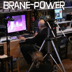 May include: A man sits at a desk in front of a computer monitor with a purple and pink screen. He is using a mouse and keyboard. There is a camera on a tripod in front of him. The text "Brane-Power" is visible above the desk.