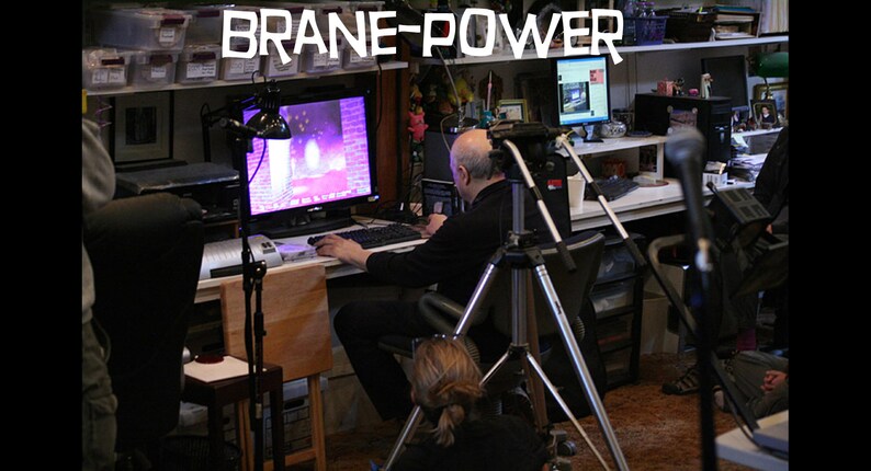 May include: A man sits at a desk in front of a computer monitor with a purple and pink screen. He is using a mouse and keyboard. There is a camera on a tripod in front of him. The text "Brane-Power" is visible above the desk.