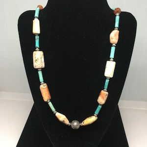 May include: A turquoise, peach, and brown stone necklace with a silver bead accent. The necklace is displayed on a black velvet stand.
