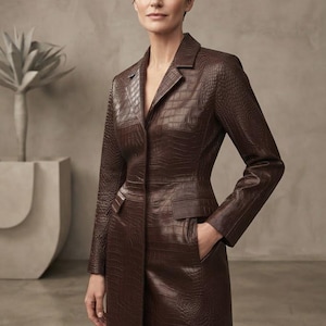 May include: A long, dark brown coat featuring a textured crocodile-embossed pattern. The coat has a structured collar, long sleeves, and two front flap pockets at the waist. The garment is fitted through the torso and extends to a full-length hemline.