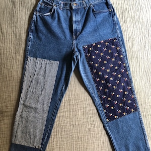 May include: Blue denim jeans with patchwork details. One leg features a vertical striped fabric panel, while the other has a floral patterned patch. The jeans have a classic five-pocket design and a button and zip closure.