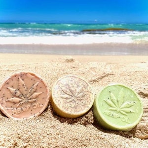 Puede incluir: Tres barras redondas de jabón con un diseño de hoja de cáñamo y las palabras "Hemp Oil" en una playa de arena.