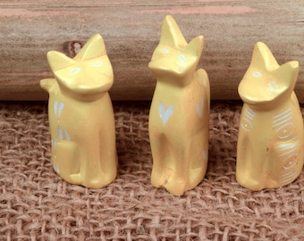 May include: Three yellow ceramic cat figurines with white painted details. The cats are standing on a brown burlap surface.