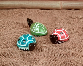 May include: Three hand-carved wooden turtles, each painted in a different color: green, blue, and red. The turtles are sitting on a brown burlap surface.