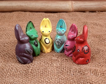 May include: A collection of six colorful rabbit figurines. Each bunny is painted in a different color, including purple, green, yellow, teal, pink, and red. The bunnies have decorative patterns and are displayed on a burlap surface.