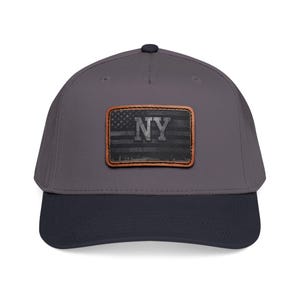 Puede incluir: Una gorra de béisbol gris carbón con visera azul marino oscuro. En el centro frontal lleva un parche rectangular con una bandera estadounidense en negro y gris, con las letras NY en blanco desgastado, bordeado en cuero marrón.