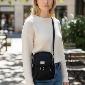 May include: Black crossbody bag with a silver-tone metal accent and a circular ring detail. The bag has a zippered main compartment and a smaller front pocket. The strap is black and adjustable. The bag is worn over the shoulder.