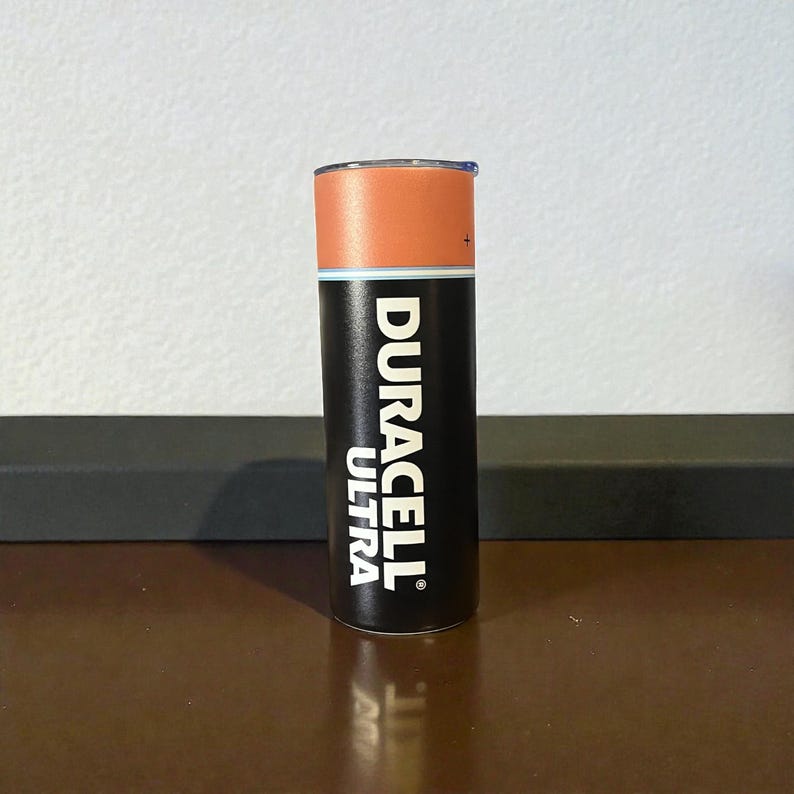 May include: A cylindrical tumbler with a design mimicking a Duracell Ultra battery. The top is orange, the middle has a white band, and the bottom is black with the Duracell Ultra logo in white. The tumbler is standing upright.