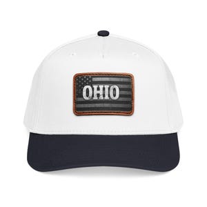 May include: A white baseball cap with a dark navy blue brim and a rectangular brown leather patch on the front. The patch features a distressed black and gray American flag design with the word OHIO printed in bold white capital letters.
