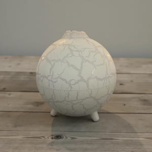 May include: A white, spherical decorative object with a cracked, gray pattern. The object has a small, textured top and rests on three short legs. The item is displayed on a wooden surface.