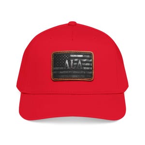 Puede incluir: Una gorra de béisbol de color rojo brillante con un parche rectangular en la parte delantera. El parche muestra un diseño de bandera estadounidense en blanco y negro desgastado con las letras ALA en el centro, bordeado por cuero cosido marrón.