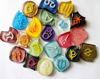 May include: A collection of colorful resin charms in various shapes, each featuring a unique symbol. The charms are in shades of blue, yellow, green, red, and purple, with glitter accents. The symbols are white or yellow, and the charms are arranged on a white surface.