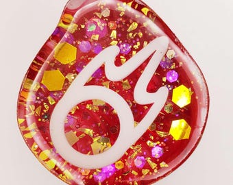 May include: A teardrop-shaped resin charm featuring a translucent red base filled with gold and purple hexagonal glitter. A white, stylized flame-like symbol is embedded in the center of the glossy, smooth surface of the decorative piece.