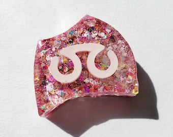 May include: A crescent-shaped resin charm filled with dense pink, magenta, and iridescent glitter. A smooth, matte, light pink symbol resembling a stylized letter or abstract icon is embedded in the center of the glittery, translucent surface.