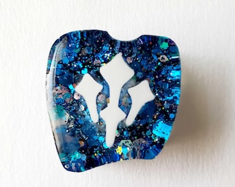 May include: A blue resin pendant with a white symbol. The pendant is filled with blue glitter and iridescent flakes. The symbol is a stylized, three-pronged design. The pendant has a rounded, irregular shape.