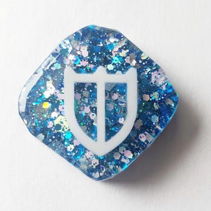 Könnte beinhalten: Ein rautenförmiger Resin-Anhänger in Blau mit Glitzer und einem weißen Schilddesign. Der Glitzer enthält blaue, silberne und irisierende Partikel. Das weiße Schildemblem befindet sich in der Mitte des Anhängers.