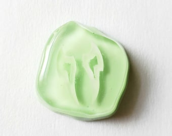 May include: A smooth, translucent light green resin cabochon with an irregular, rounded shape. The center features a recessed, white, abstract graphic design that resembles two symmetrical, curved, pointed shapes facing each other.