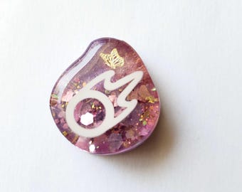 May include: A teardrop-shaped resin charm in shades of purple and pink, featuring a white flame-like graphic, gold glitter flakes, and a small gold butterfly decal embedded within the smooth, glossy surface of the decorative piece.