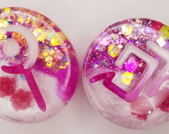 May include: A pair of circular resin ear plugs featuring a translucent pink and white gradient base with embedded holographic glitter and small pink floral accents. Each plug displays a distinct white raised symbol on the front surface.