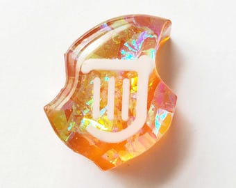 May include: A translucent orange resin charm with a white symbol. The charm has an iridescent effect, with flashes of blue, green, and yellow. The white symbol is a stylized design with a curved top and three vertical lines.