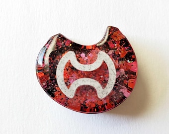 May include: A resin pendant with a white, speckled, double-curve design on a background of red and black glitter. The pendant has a rounded shape with a small notch at the top.