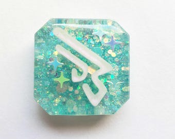 May include: A square, teal-colored resin piece with glitter and star-shaped accents. A white, stylized design is embedded in the center. The resin has a slightly iridescent quality, catching the light. The overall effect is whimsical and decorative.