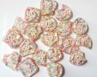 May include: A collection of small, clear resin charms in various geometric and organic shapes, each embedded with a dense mix of multicolored confetti-like glitter and featuring a single white embossed symbol on the front surface.