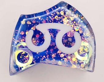 May include: A glossy, irregular-shaped blue resin charm featuring a white symbol resembling a stylized omega or double loop. The surface is embedded with iridescent hexagonal glitter and two circular, metallic-toned rings visible inside.