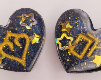 May include: Two heart-shaped objects with a dark blue, glittery base. Each heart features gold-colored symbols and small, iridescent star-shaped accents embedded within a smooth, glossy finish. The design includes a mix of gold glitter and holographic star elements that catch the light, creating a reflective surface across both heart-shaped pieces.