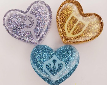 May include: Three heart-shaped resin charms with glitter. One is purple with a staff symbol, one is gold with a shield symbol, and one is blue with a crest symbol. Each charm has a smooth, glossy finish and a distinct, centered light-colored icon.