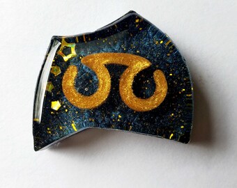 May include: A resin-cast accessory with a dark blue background speckled with gold glitter. A gold-colored symbol is centered on the accessory. The edges are clear and slightly curved.