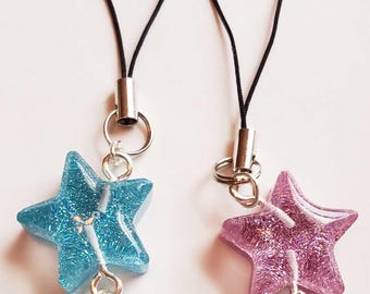 May include: Two phone charms featuring glittery resin star and heart pendants. One set is blue with a white symbol on the heart, and the other is pink with a white symbol on the heart. Each charm hangs from a black cord with a silver metal connector.