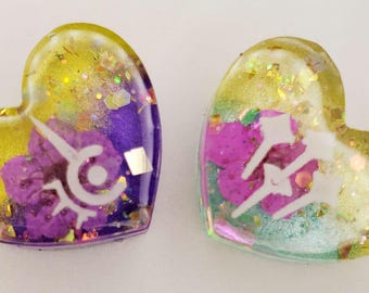 May include: Two heart-shaped resin charms featuring a glossy finish with embedded gold glitter and iridescent flakes. The left charm contains purple floral elements and a white symbol, while the right charm displays three white wand icons.
