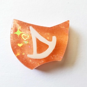 May include: Orange resin charm with a white letter "D" and glitter accents. The charm has a unique shape with a small hole at the top. Decorative hearts and stars are embedded in the resin.