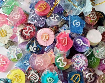 May include: A collection of colorful resin charms with glitter and various symbols. The charms are in various shapes, including circles, hearts, and irregular forms. Some charms have gold accents and are scattered with small organza bags.