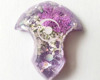 May include: A shield-shaped resin pendant in a translucent purple hue, embedded with dried purple and white flowers, iridescent glitter flakes, and a central white circular emblem featuring a stylized crescent moon and dot design.