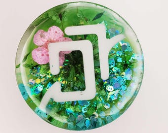 May include: A circular resin piece featuring a translucent green base embedded with dried green foliage, a small pink flower, and iridescent blue hexagonal glitter. A white, stylized, angular graphic symbol is layered over the center.